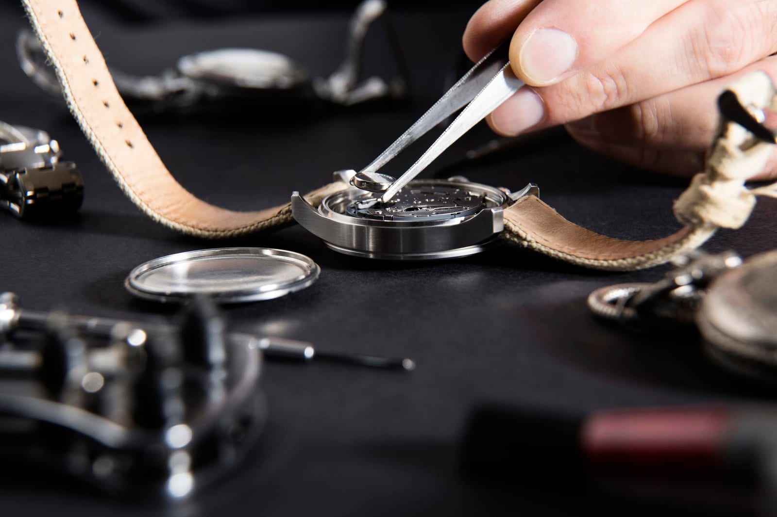 Mani con pinzette che richiudono la cassa di un orologio, con cinturini in pelle sul banco