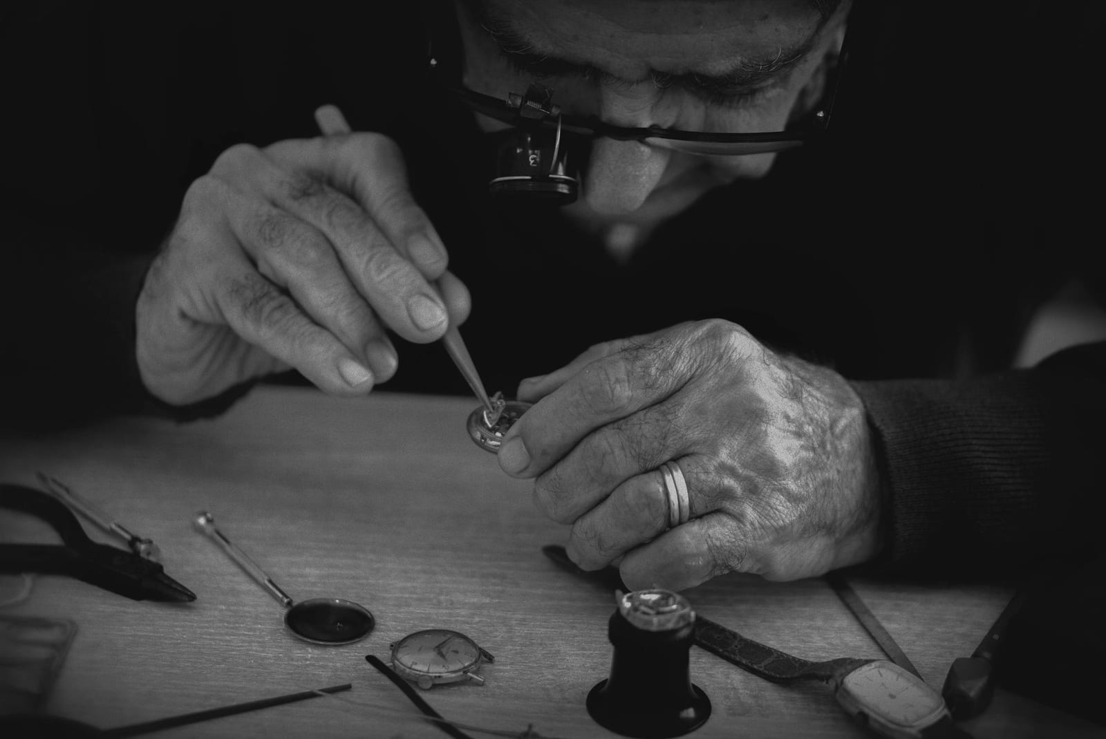 Ritratto in bianco e nero di un orologiaio al lavoro con lente da occhio, strumenti e componenti sul banco